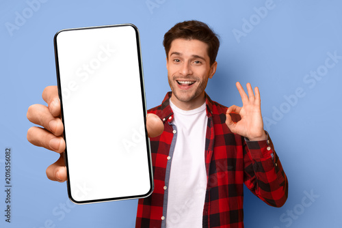 Fotografie A man stands in front of a blue wall holding a smartphone with a blank screen