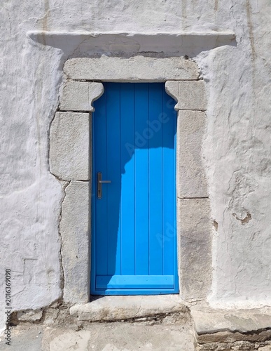 Konstfotografi Photo of a traditional cobalt-blue wooden door set into a sunlit whitewashed wall, capturing a charming Cycladic architectural detail on Mykonos Island, Greece