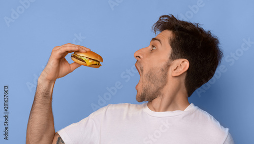 Canvastavla A man stands against a blue background holding a burger in one hand while opening his mouth wide