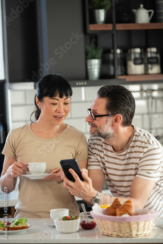 Couple having breakfast in modern kitchen while checking smartphone together. Asian woman holding coffee cup and Caucasian man using mobile phone during relaxed morning routine