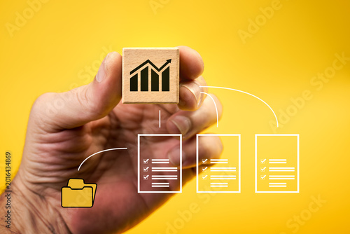 A hand holds a wooden block with a graphic symbol of clarity and growth in business on a yellow background. Arrows connect the block to documents and folders. Concept of data, reports, and business or