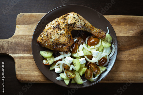 Grilled chicken leg rests on a dark plate. Cucumber, tomatoes, and onions create a fresh contrast. Wooden cutting board adds rustic charm. Warm tones suggest a hearty, home-cooked meal