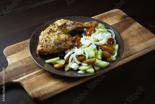 Grilled chicken leg rests on a dark plate. Cucumber and cherry tomatoes garnish the dish. Wooden cutting board provides rustic contrast. Warm lighting enhances meal's inviting appeal