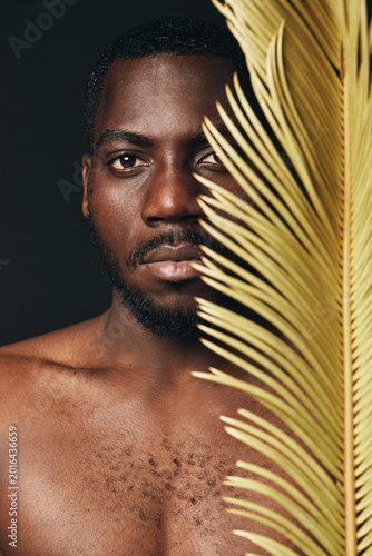 Foto African man with dark skin and beard in creative conceptual visual style, half face covered by yellow palm leaf, closeup portrait with artistic lighting and natural texture on black background