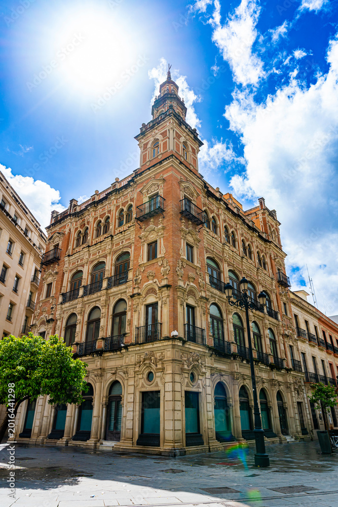 Fototapeta premium Beautiful historic building with richly decorated facade and elegant tower on a street corner in Seville. Urban scene with people walking, bright sunlight and blue sky in Spain.