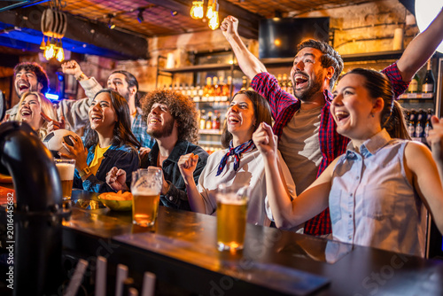 Excited friends shouting and celebrating soccer goal with beer in a pub.