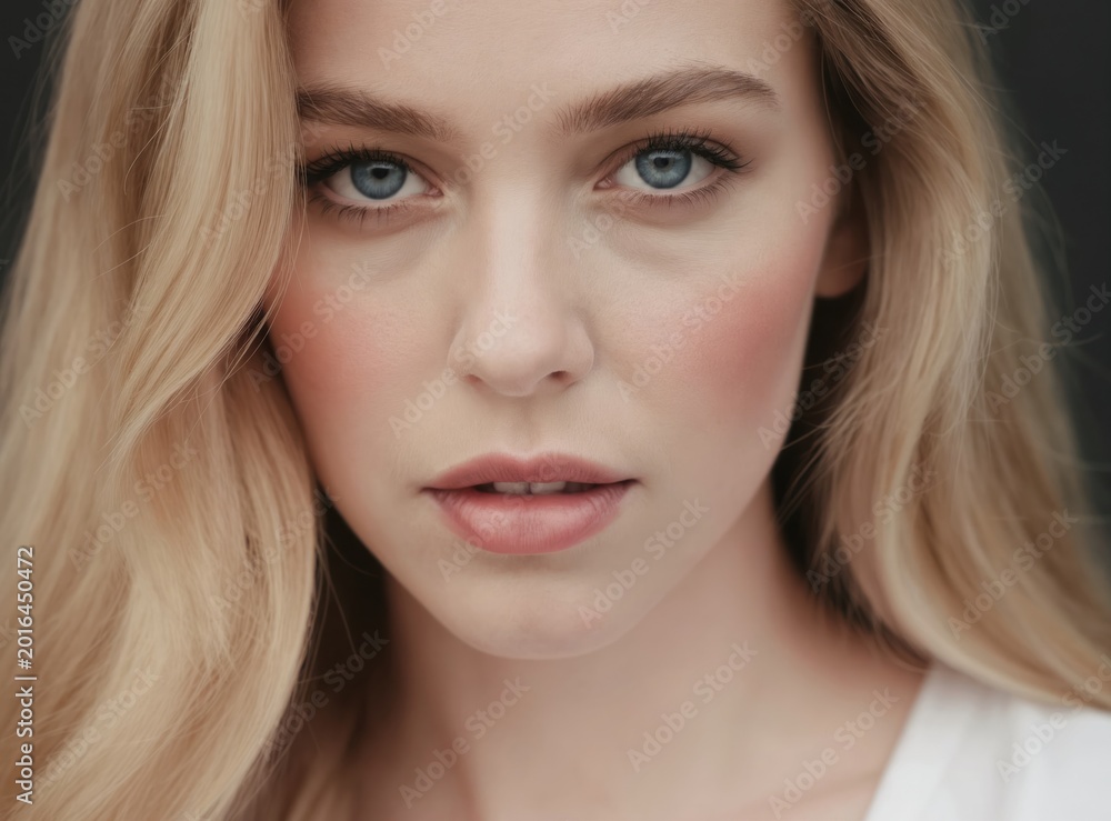 Naklejka premium Close up portrait of a young blonde woman with blue eyes. She has clear skin and light makeup with blush. Her hair is wavy and blonde. She wears a white shirt.