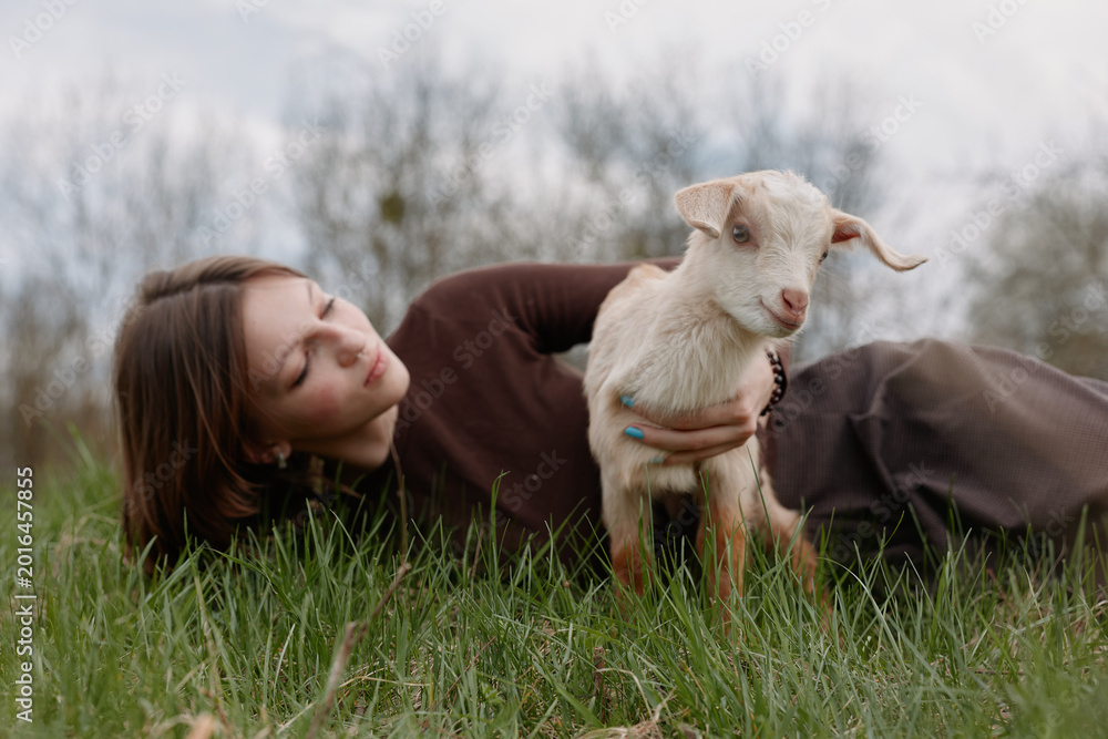 Obraz premium Lifestyle scene with a woman lying on lush grass, a goat kid nearby, soft light, candid expression, natural colors, peaceful outdoors, gentle affection and connection in a rural meadow