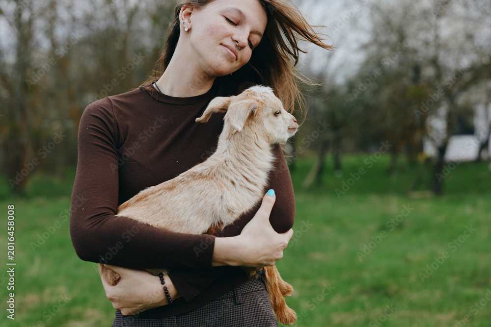 Fototapeta premium Joyful woman outdoors holding a goat kid in her arms, park setting, casual sweater, candid lifestyle moment, natural light, warm mood, gentle bond with animal, authentic and serene countryside scene