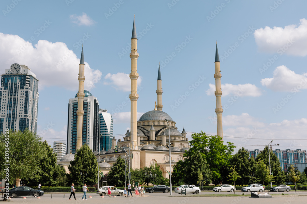 Fototapeta premium Wide view of the Heart of Chechnya Mosque named after Akhmad Kadyrov, with tall minarets, trees, cars and pedestrians in Grozny, Chechnya, Russia