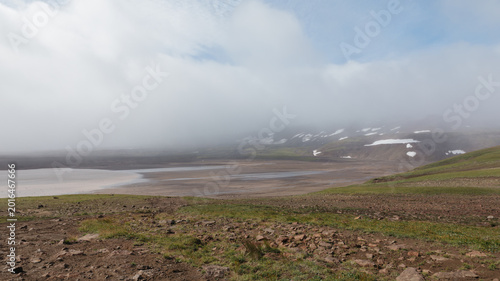 Wide view of a remote volcanic lake with stony tundra slopes and low clouds drifting over the hills, shot in Kamchatka, Russia, in calm overcast weather