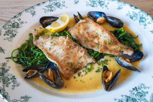 Pan fried fillet of plaice served with samphire, spinach and mussels in a butter sauce. The plate is large and white green decoration on a wooden table.