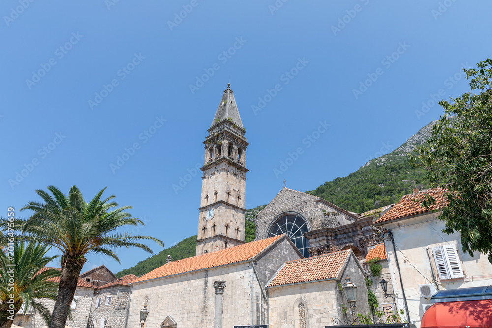 Obraz premium Sunny view of the historic church bell tower and stone buildings with red tile roofs in Perast, Kotor Bay, Montenegro, with green mountains and clear blue sky
