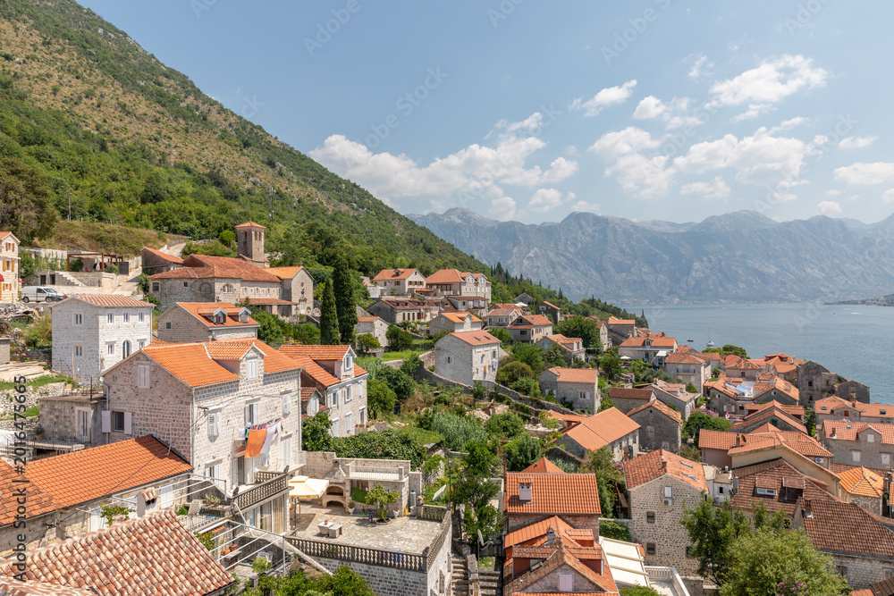 Fototapeta premium Elevated panorama of Perast with stone houses and red tile roofs above the Bay of Kotor, backed by steep mountains and blue sea under a sunny sky, Montenegro