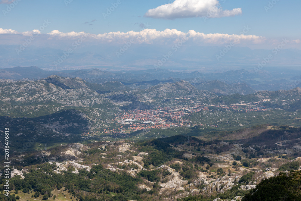 Obraz premium Wide panoramic view from Lovcen National Park overlooking the town of Cetinje, Montenegro, with rugged karst mountains, green forests, distant haze and clouds