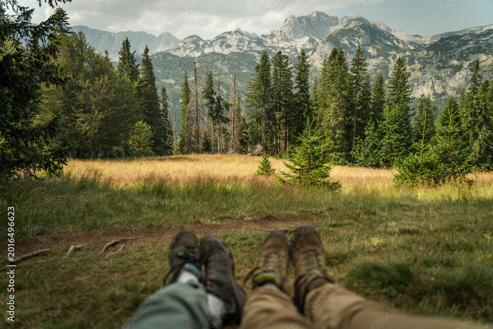 Fototapeta premium Panoramic view of high mountains with blurred legs of hikers in the foreground sitting on a green meadow, selective focus.