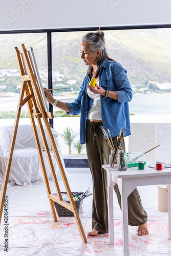 Middle-aged painter painting canvas at easel, holding yellow jar and brush, denim barefoot studio