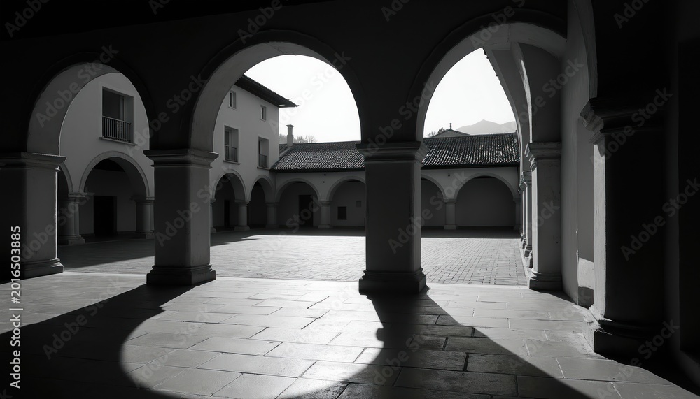 Fototapeta premium Monochrome building facade with arched walkways forming courtyard. Dramatic light, shadow play across paving stones, colonnade. Geometric patterns, architectural details create classic, serene