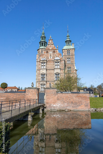 A stunning front view of Rosenborg Castle in Copenhagen, Denmark, with beautiful reflections in the surrounding moat under a clear blue sky.