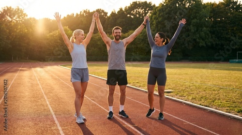 Three smiling athletes stand together on a red running track, raising their hands in a joyful victory.