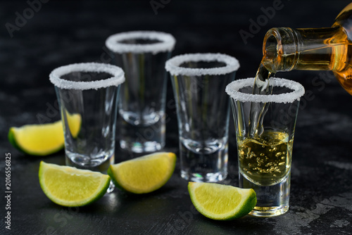 pouring golden tequila into a glass with lime slices and salt crystals on a dark table, selective focus.