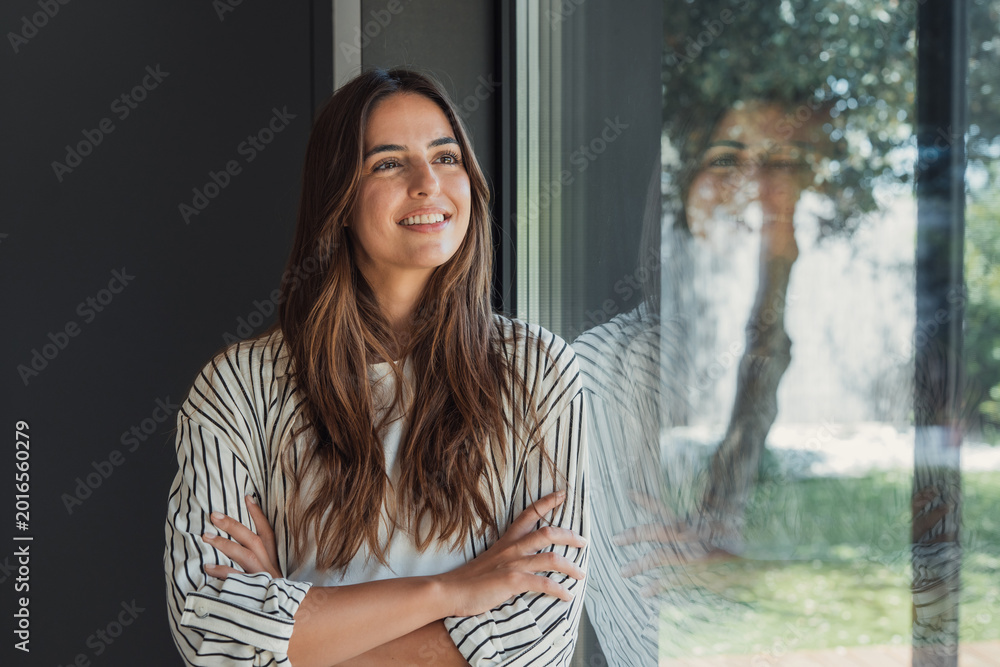 Fototapeta premium Cheerful young woman standing indoors with arms crossed, smiling as she looks out a large window, reflecting a blurred garden with trees and natural daylight
