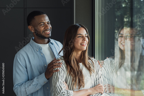 Tableau sur toile Diverse couple standing by a window, smiling and embracing as they look toward t