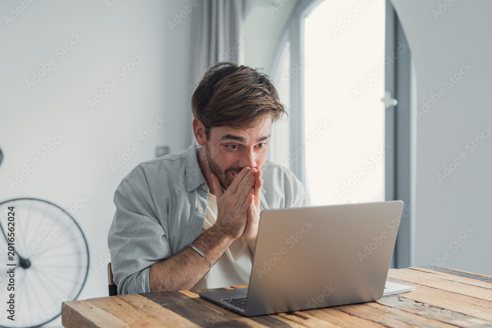 Fototapeta premium Young man reacting to surprising news or unexpected information on his laptop, expressing disbelief and wonder while sitting at a wooden desk in a bright room