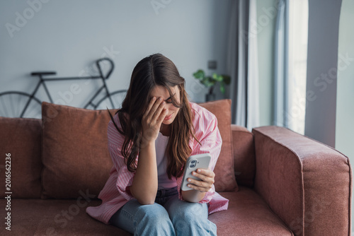 Young woman sitting on a sofa at home, crying and covering her face with one hand while holding a mobile phone in the other, expressing stress, sadness, and emotional distress over bad news