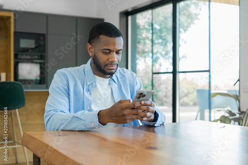 Konstfotografi Black man sitting at a wooden table in a modern home, focused on scrolling and i