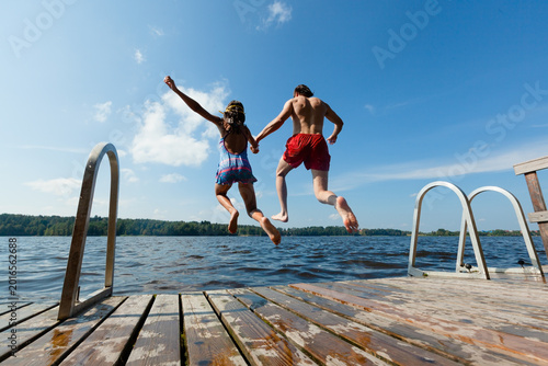 Man and girl jump off dock into lake in extreme summer fun on sunny day. Guy in red trunks and girl leap together in thrilling outdoor adventure. Exciting duo creates splash with daring jump