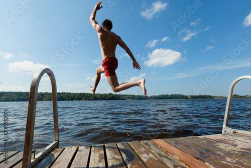 Bilde på lerret Guy jumps off dock captured in midair flight over lake on sunny summer day