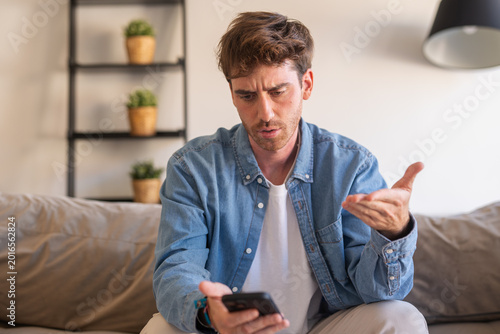 Bilde på lerret Young man on a sofa reacts with confusion and frustration to bad news on his sma