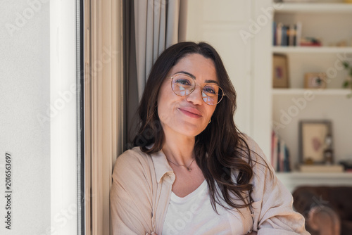 Obraz na plátně Cheerful young adult woman with brown hair and glasses leaning against a window