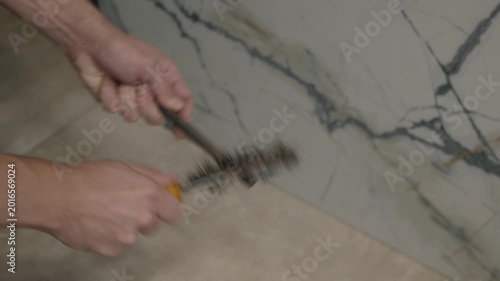 Hands cleaning metal pipe with wire brush, bristles scraping rust and debris against marble wall, steady manual strokes, particles falling, focused restoration and plumbing maintenance in workshop