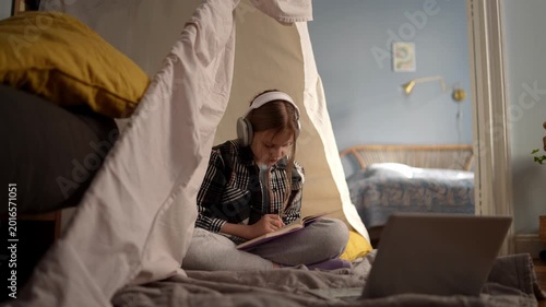 White girl with headphones in fort, sitting crosslegged with laptop and notebook, glowing screen and ambient lamp light, notetaking during online lesson, .