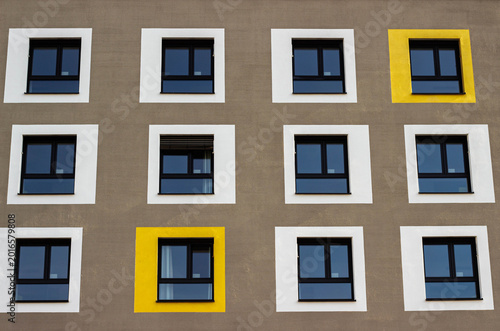 Modern building facade featuring a grid of rectangular windows with contrasting yellow and white frames against a textured gray wall in an urban setting