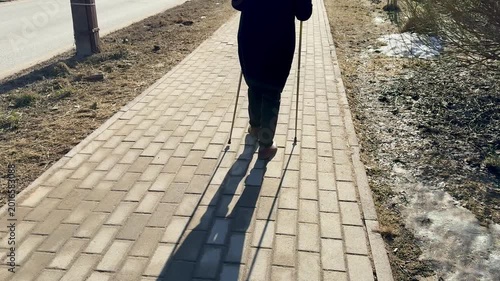 Woman Nordic walking shaded park path, brisk stride with poles and long shadow cast on paving, lea strewn verge, cool crisp light