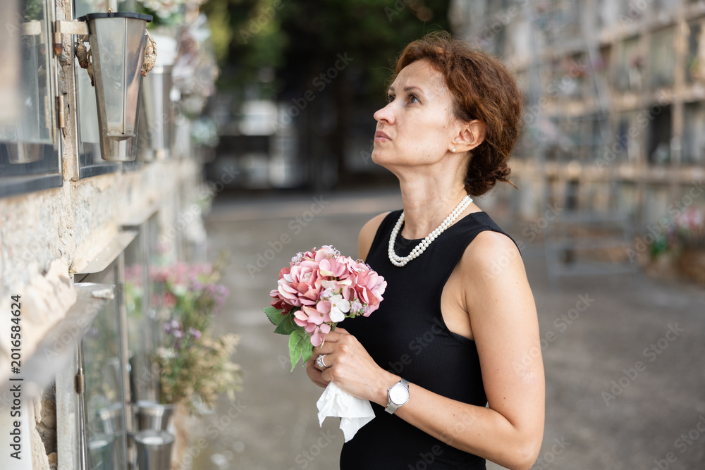 Fototapeta premium Sorrowful middle aged woman in black dress standing in cemetery holding bouquet of flowers with reflective and somber expression in front of stone memorial wall
