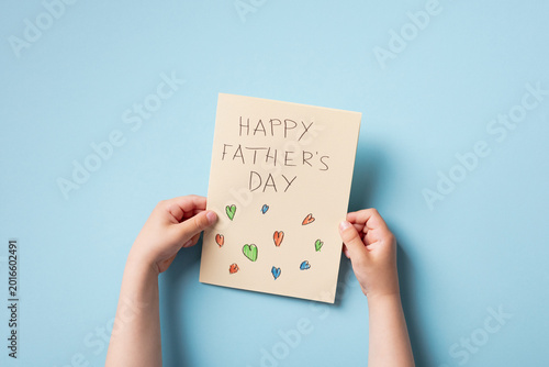 Top view of a child's hands holding a handmade Father's Day card on a blue background.