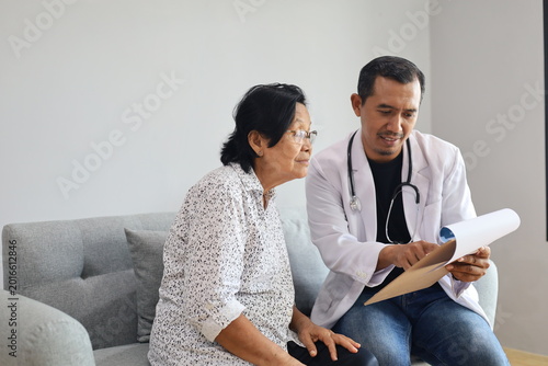 Asian male doctor talking with elderly senior woman in his home. Mature woman patient and medical worker in uniform holding clipboard noting personal information