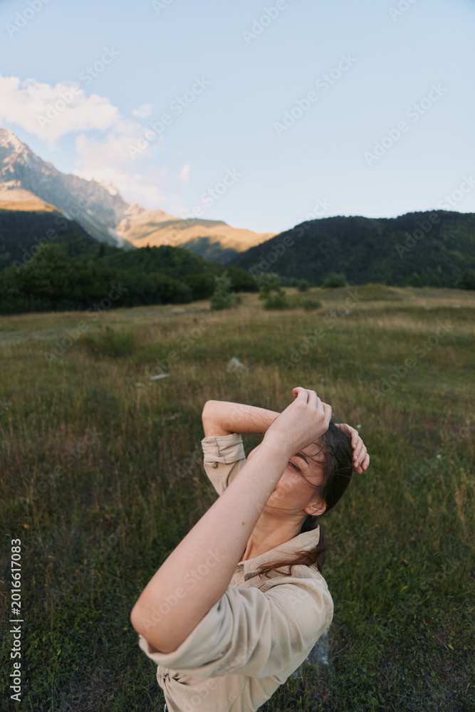 Naklejka premium Young woman in a light jacket stands in a grassy field with distant mountains, arms raised, enjoying fresh air and warm sunlight in nature.