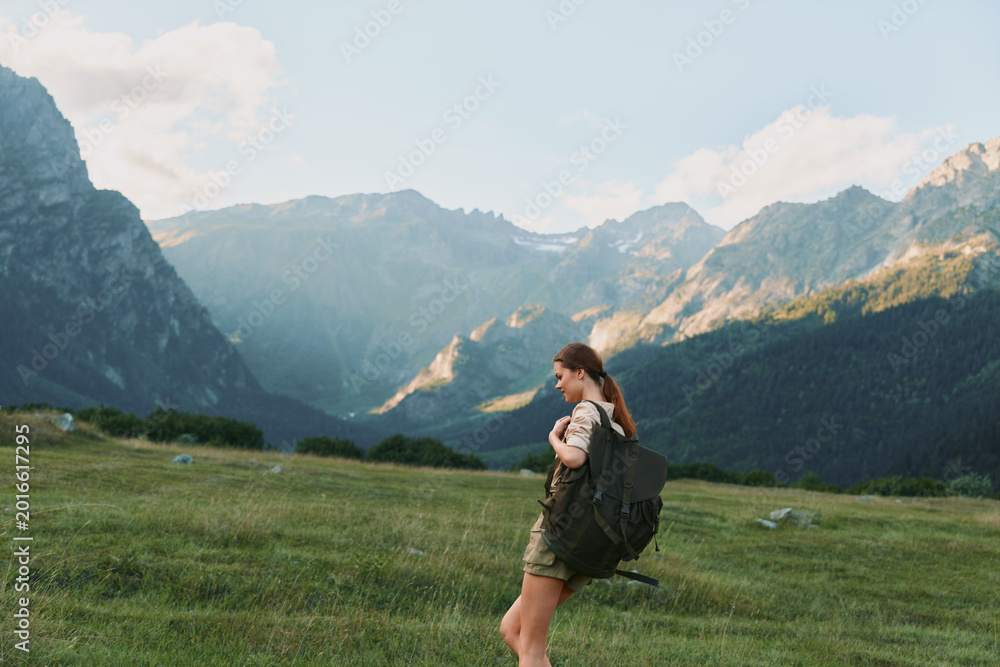 Fototapeta premium Hiker with a large backpack strides across a sunlit alpine meadow under towering mountains, embodying freedom and adventure in a single warm moment