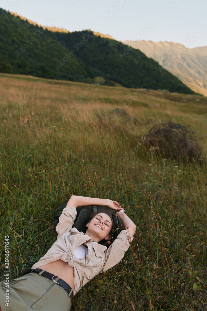 Naklejka premium A woman lies in a grassy meadow, arms folded behind her head, smiling softly. Broad hills and warm light create a serene outdoor lifestyle portrait today
