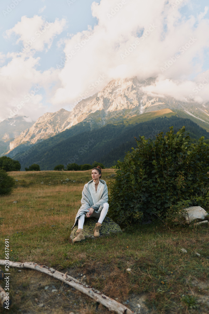 Naklejka premium Young woman in casual clothes sitting on a rock in a mountain meadow with cloudy sky. Concept of lifestyle, nature, and outdoor relaxation in fresh air.