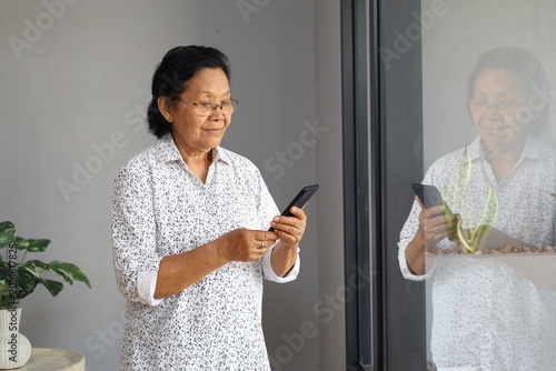 Happy senior Asian woman standing by window, holding cellphone in hands, elderly lady contacts her family