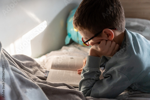 Curious boy in glasses lying in bed reading book under warm sunlight at home. Concept of childhood education, love of reading and cozy home leisure lifestyle. High quality photo