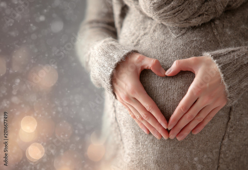 Close up of pregnant belly with hands forming heart shape, showcasing warmth and love. soft sweater adds cozy touch, while blurred background creates dreamy atmosphere