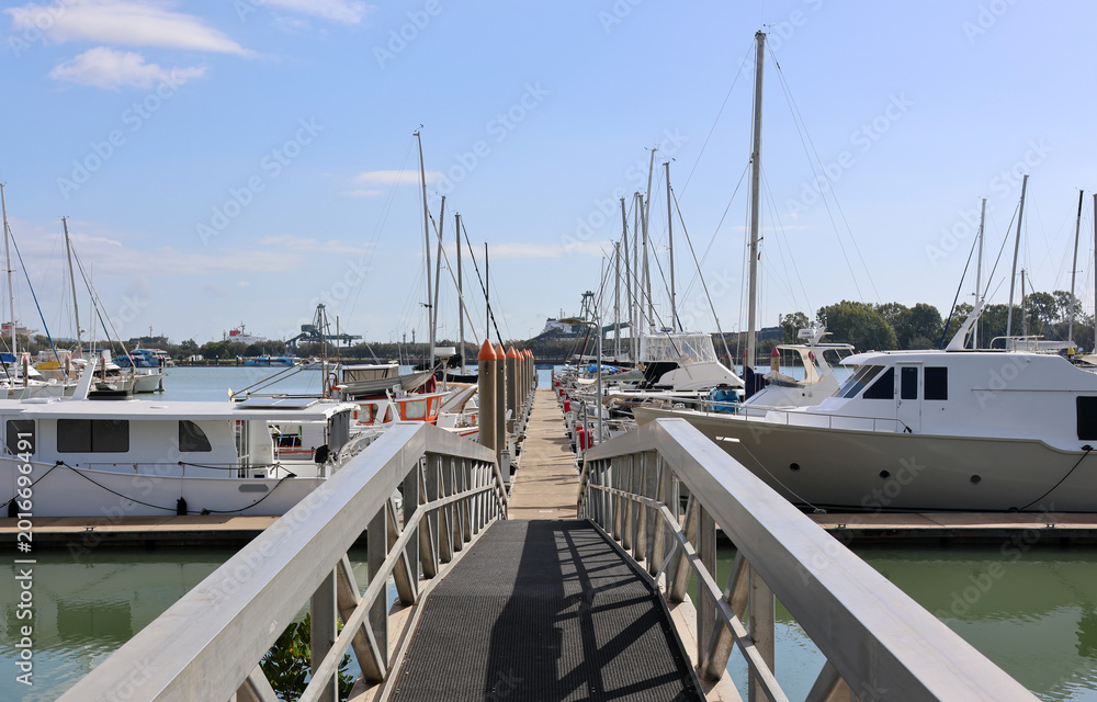 Fototapeta premium Metal gangway leading to a long pontoon lined with motorboats and yachts at the Gladstone Marina under a clear blue sky in Queensland, Australia