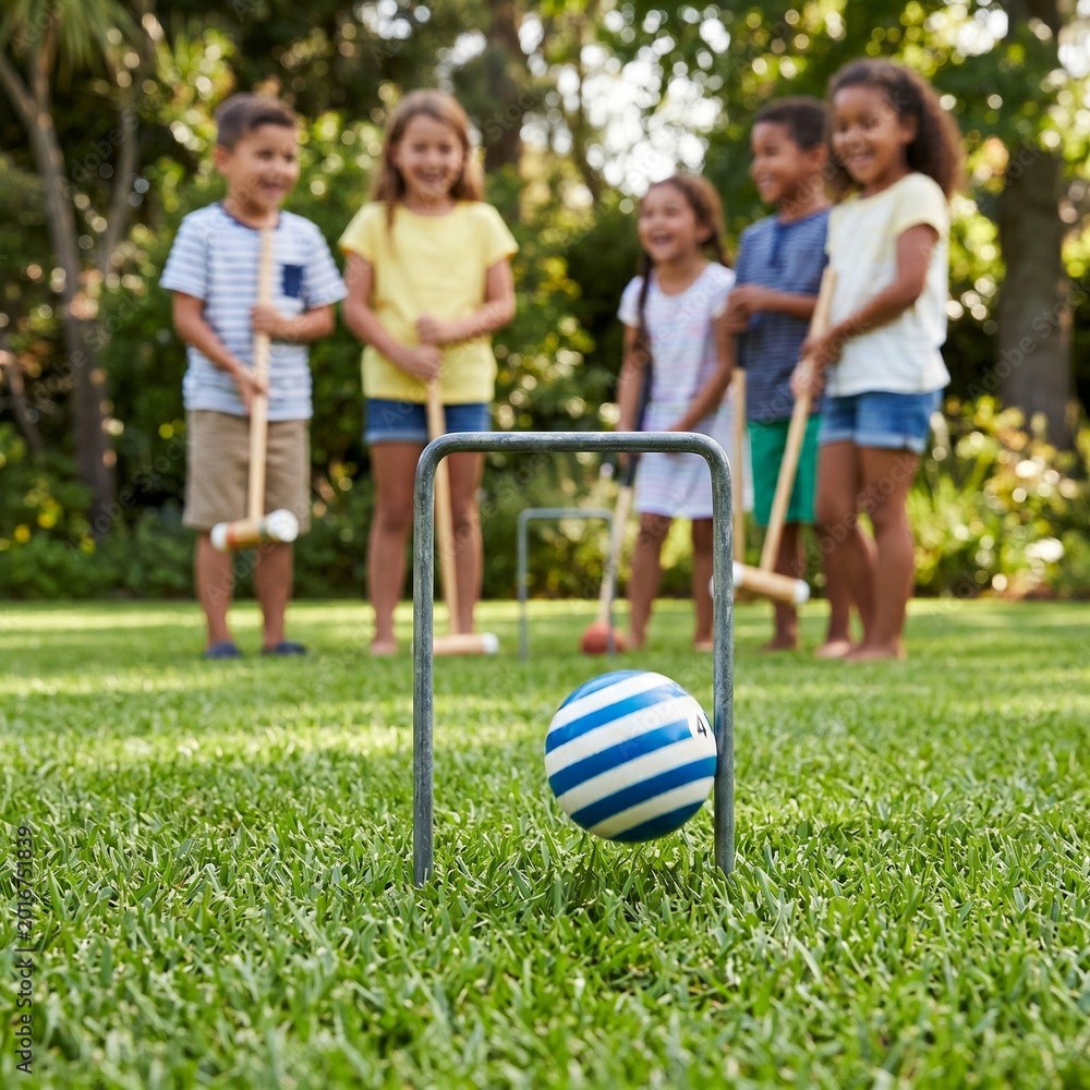Obraz premium A striped croquet ball passing through a metal wicket on a manicured lawn, with children holding mallets in the blurred background, highlighting youthful recreation 1:1 ratio.